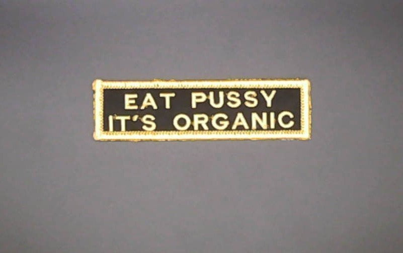 A rectangular embroidered patch with a black background and yellow border, featuring the text 'EAT PUSSY IT'S ORGANIC' in yellow lettering, shown against a solid gray background.