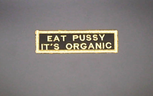 A rectangular embroidered patch with a black background and yellow border, featuring the text 'EAT PUSSY IT'S ORGANIC' in yellow lettering, shown against a solid gray background.
